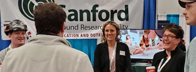 Educators and a researcher with the South Dakota Science and Technology Authority (SDSTA) at SURF speak with conference participants in Denver during the 2024 National Science Teaching Association Conference.  Poeple laughing and smiling while taking with each other at the SURF Booth at the conference. 