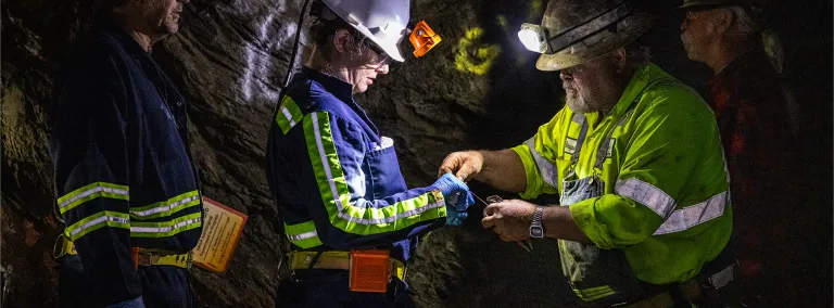 Two former Homestake workers, Al Pfarr (right) and Bryan Silvernail working with scientists Magdalena Osburn and Bradley Stevenson with the Deep Mine Microbial Observatory (DeMMO) to repair a biologic sampling site on the 800 Level of SURF.  The photos is in a dark area, surrounded by rock inside the mine, individuals are wearing PPE and bright reflective clothing used underground. 