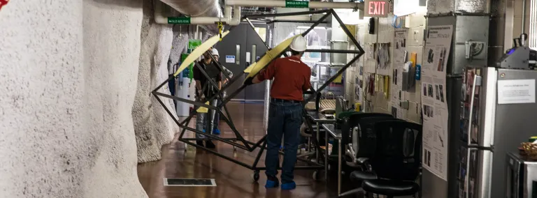 Two people move a large metal framed box through a tight area in an underground lab.