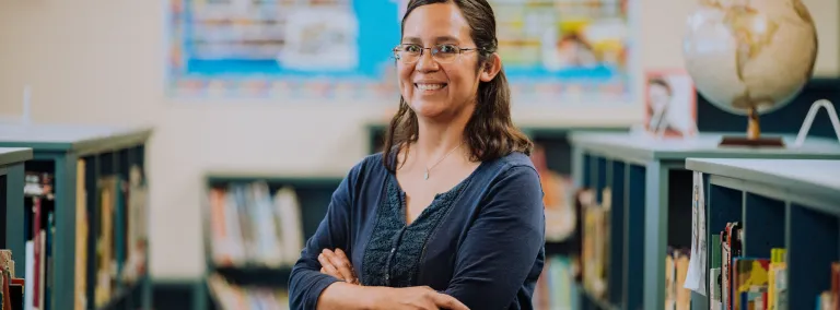 a person stands with their arms crossed and smiles amidst bookshelves