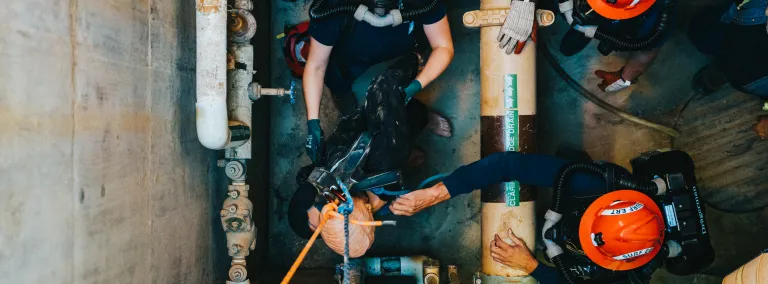 a person wearing a full-face breathing apparatus looks up through a utility hole as the emergency rescue team raises a training mannequin using ropes.