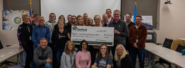 In a conference, a group of 20 people stand together around a large check made out to the Foundation for Health 