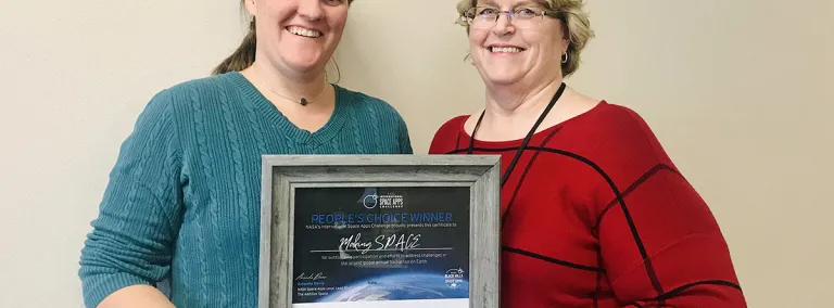 Natasha Wheeler and Caroline Jones stand against a white wall, holding a framed certificate of their global recognition 