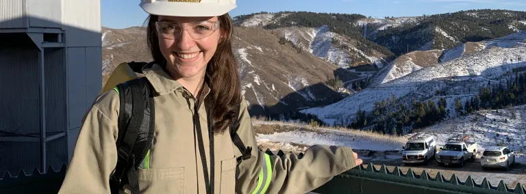 Researcher in hardhat and safety glasses stands in front of hilly landscape