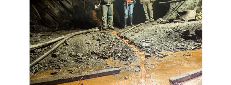 Three people stand in front of stream of brown water that is flowing into a borehole.