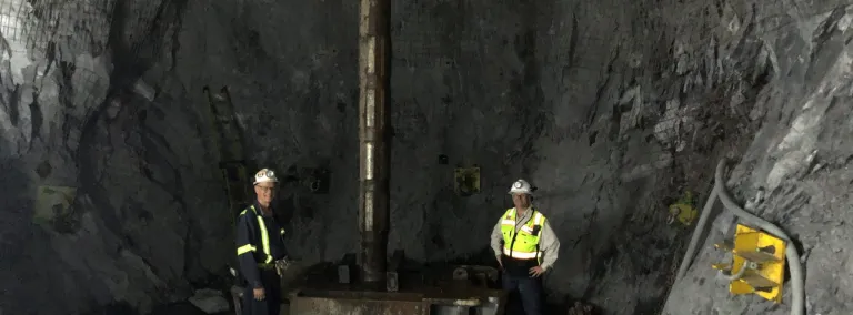 Fermilab LBNF engineers James Rickard and Syd Devries stand beside the 12-foot reamer at the 4,850-foot level, showing the enormous size of the reamer bit used to drill the ventilation shaft. 