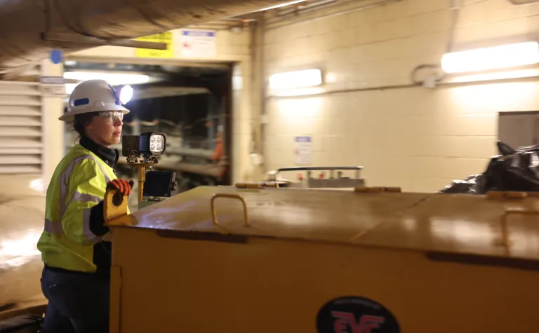 Sarah Wortman, an underground facilities technician at SURF,  dressed in a hardhat, light, and reflective vest with red gloved, operates a locomotive on the 4850 Level undergroudn at SURF.