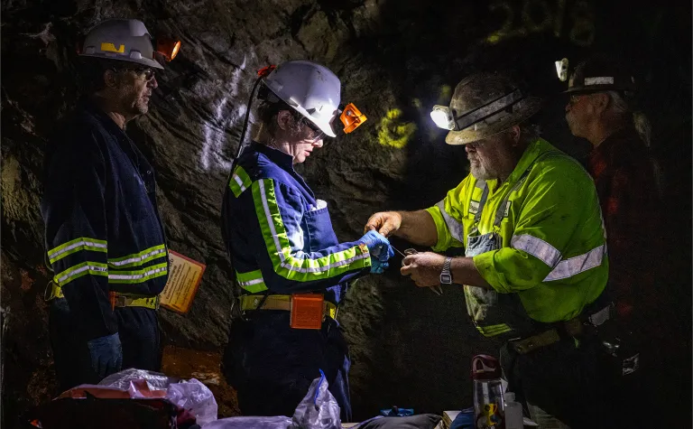 Two former Homestake workers, Al Pfarr (right) and Bryan Silvernail working with scientists Magdalena Osburn and Bradley Stevenson with the Deep Mine Microbial Observatory (DeMMO) to repair a biologic sampling site on the 800 Level of SURF.  The photos is in a dark area, surrounded by rock inside the mine, individuals are wearing PPE and bright reflective clothing used underground. 