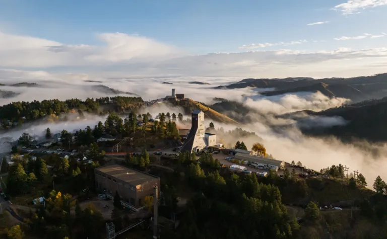 The Ross and Yates headframes, at the Sanford Underground Research Facility, sit above the clouds covering the Black Hills in October of 2023. This image of fog rolling in between the Ross and Yates headframes at the Sanford Underground Research Facility (SURF) during a sunrise in October of 2023. In rare cases, fog will cover the lower lying valleys and hills around SURF, leaving the town of Lead exposed in the sunlight, above the clouds, like an island on the ocean.