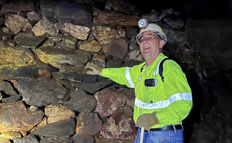 Kyle Ehnes  underground at SURF wearing a reflective shirt and proper PPE smiling with his had on a rock wall. 