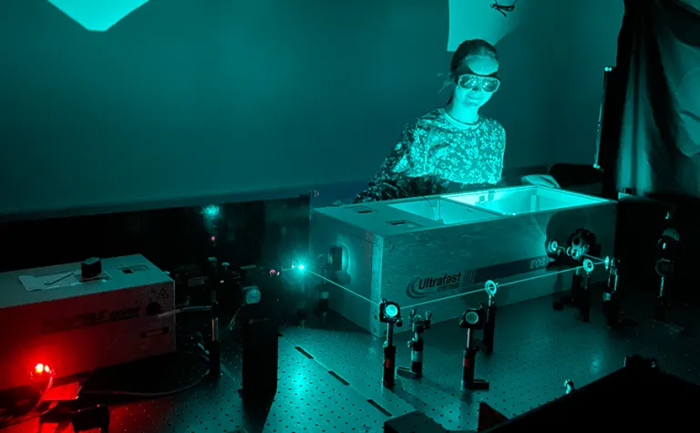 Madison Jilek works in her laboratory at the University of Colorado Boulder.  She is wearing laser protective eyeware, in a room illuminated by green light with a green colored laser running through a series of lenses and mirrors around a table. 