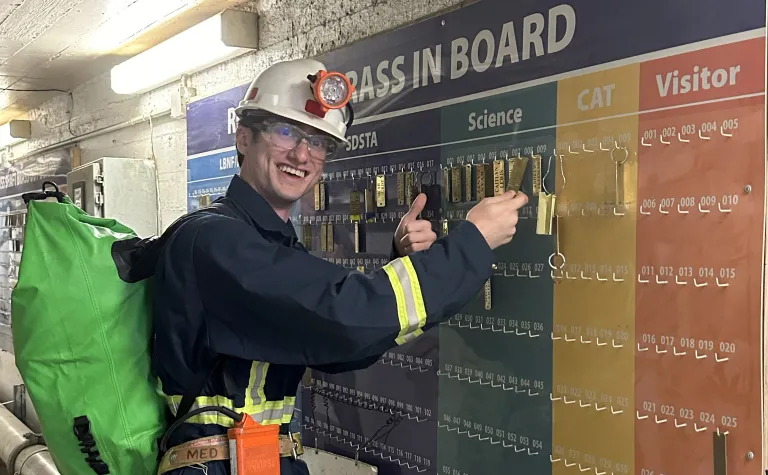 Archer Sagaskie, a student of physics at Grand Valley State University in Michigan smiles as he places a brass tag on the board before going underground at SURF.  Archer is in full SURF PPE, including a hard hat, overalls, safety glasses, and a W65 resperator on his belt.