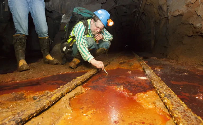 Dr. Dave Bergmann takes a sample from an underground area at SURF in between two train rails in rust colored fluid