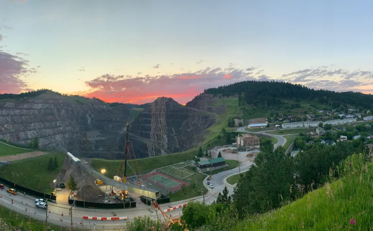 A crane lifts a large segment of steel conveyor truss in the air, over the highway. In the background, there is an open pit in the earth, created by strip mining. 