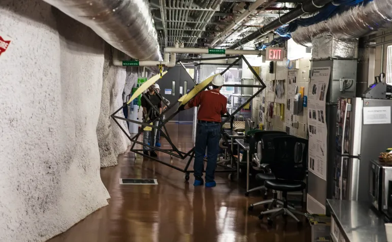 Two people move a large metal framed box through a tight area in an underground lab.