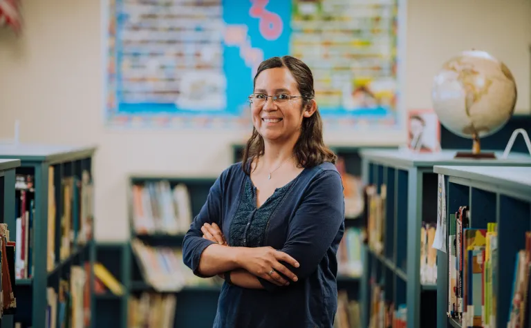 a person stands with their arms crossed and smiles amidst bookshelves