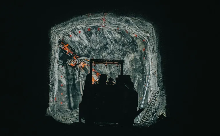 The silhouette of drilling machine stands out against a well-lit rock wall.  