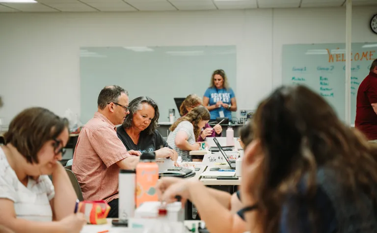 Two educators sit at a table, looking at paper materials, in a room full of other educators during a professional development workshop 