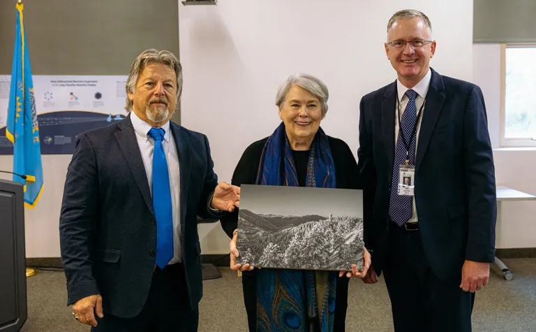 Three people stand facing the camera, the person in the middle holds a photo 