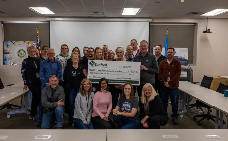 In a conference, a group of 20 people stand together around a large check made out to the Foundation for Health 