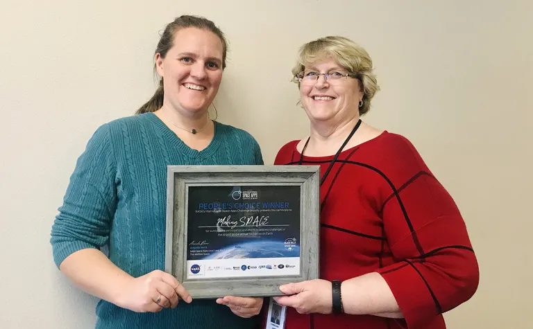 Natasha Wheeler and Caroline Jones stand against a white wall, holding a framed certificate of their global recognition 