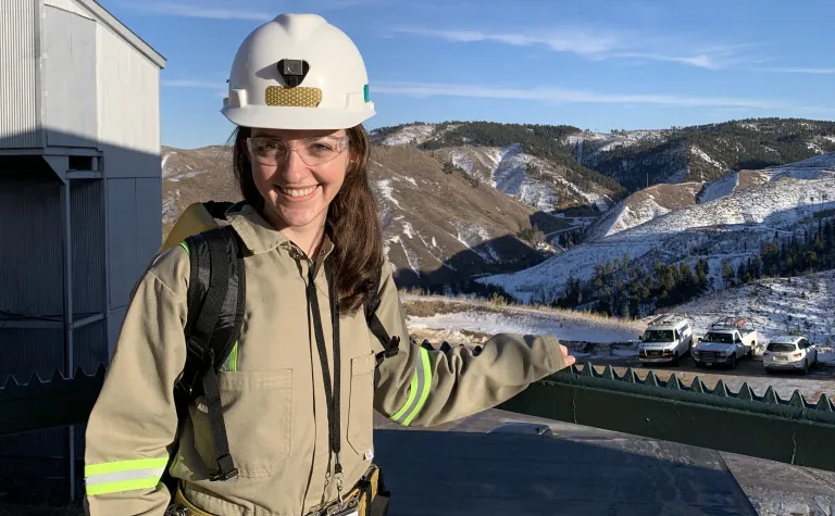 Researcher in hardhat and safety glasses stands in front of hilly landscape