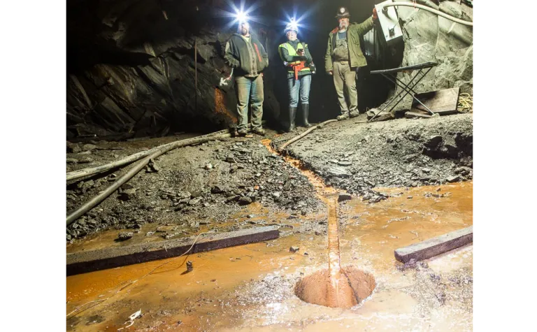 Three people stand in front of stream of brown water that is flowing into a borehole.