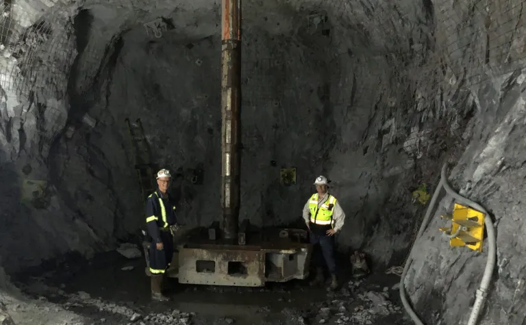 Fermilab LBNF engineers James Rickard and Syd Devries stand beside the 12-foot reamer at the 4,850-foot level, showing the enormous size of the reamer bit used to drill the ventilation shaft. 