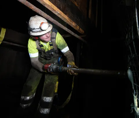 Will Hover, an infrastructure technician at SURF, tightens a bolt inside the Yates Shaft. Will is in full PPE holding a large red pneumatic driver with a long socket made for tightenign rock bolts.  It's dark and he is crouched in a small confined spaqce under some timber and pipes above him. Photo by Adam Gomez.