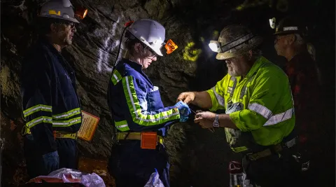 Two former Homestake workers, Al Pfarr (right) and Bryan Silvernail working with scientists Magdalena Osburn and Bradley Stevenson with the Deep Mine Microbial Observatory (DeMMO) to repair a biologic sampling site on the 800 Level of SURF.  The photos is in a dark area, surrounded by rock inside the mine, individuals are wearing PPE and bright reflective clothing used underground. 