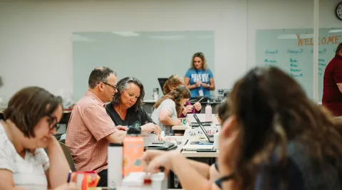 Two educators sit at a table, looking at paper materials, in a room full of other educators during a professional development workshop 